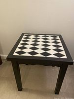Square table with black and white tile top in diamond pattern, black legs and base visible against a carpeted floor and light colored wall.