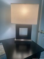 Photo of black and silver geometric base table lamp with a white rectangular fabric shade on a black surface.