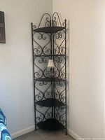 Black metal wrought iron corner shelf unit photographed in a room corner with a small lamp on one shelf. Shows full height and width, and decorative scrollwork details.