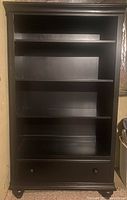 Front view of black solid wood shelving unit showing four shelves and a drawer at the bottom.