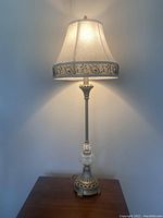 Full view of the tall table lamp lit on, showing antique gold and silver metal base with clear glass accent and white fabric damask conical shade trimmed with embroidered black floral design