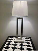 Tall silver lamp with open rectangular metal frame, white marble base, and white rectangular fabric lampshade against a neutral wall.
