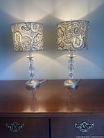 Pair of lamps shown on wooden surface, displaying brushed nickel bases with clear crystal stacked segments and black and white paisley fabric shades, both lit emitting soft light on wall.