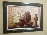 Front view of framed wall art showing an image of a composition of urns, moss balls, and vases in warm neutral colors, in a carved black frame with cream inner border.