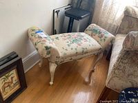 Full view of bench showing floral ivy upholstery, curved arms, white painted wooden base and legs, placed on hardwood floor next to a floral sofa.