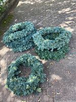 Three green artificial Christmas wreaths placed on an outdoor paved area, showing needles and integrated string lights.