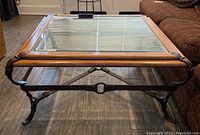 Full view of square coffee table on wood floor showing glass top, wooden frame and metal legs