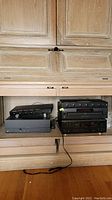 Photo of black audio equipment including two Niles 6-speaker control systems stacked on the right and a Denon 5-disc changer with a Sonance amplifier to the left, all set inside a wooden cabinet.