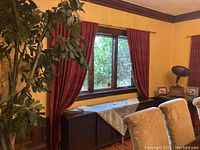 Room view showing one pair of red patterned drapes hanging beside a window above a cabinet with plants and furniture visible.