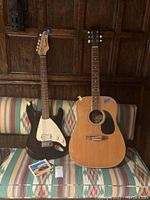 First Act electric guitar and acoustic guitar sitting on a patterned sofa with box of guitar strings in front.