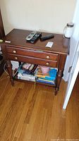Front view of one Ethan Allen wooden nightstand showing two drawers with brass knobs and lower shelf holding books and other items. Wood shows minor scratches on top.
