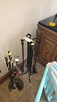 Photo showing both sets of fireplace tools side by side placed on tile floor near wooden cabinet, clear view of tools and stands