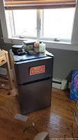 Front view of the Haier mini fridge showing stainless steel body with black top and handles, placed on hardwood floor.
