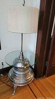 Full view of the lamp table showing glass top, metal base with concentric circular patterns, and light drum lamp shade.