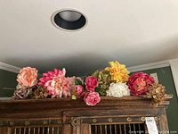 Wide view showing assortment of silk faux flowers and planter on top of wooden furniture against wall.