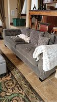 Full view of gray upholstered sofa with nail head trim, multiple matching pillows and a white throw blanket, shown with tile floor and wooden chairs in the background.