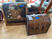 Two storage chests placed on a floor, one black with detailed floral painting and brass rivets, the other smaller brown with rivet detailing and visible wear.