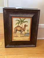 Framed art print of a camel standing near a small pond with palm trees in the background, dark brown textured wide frame.