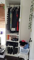 Front view of white wardrobe cabinet showing clothes on hangers and three open shelves with shoes and books.
