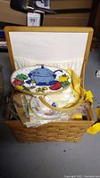 Wicker sewing basket open showing cross stitch fabric and supplies inside including an oval cross stitch piece with kitchen items.