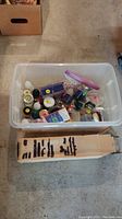 Plastic bin filled with various jars and bottles containing beads and jewelry findings placed on a floor surface.