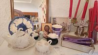 White ceramic pumpkin teapot, blue and white plate, various candles and candleholders arranged on a surface with a mirror in the background.