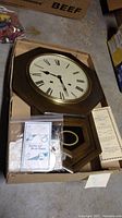 Wooden octagonal framed wall clock with brass rim, white face with Roman numerals lying in a cardboard box with small bag containing repair or winding accessories.