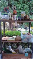 Full view of three-tiered metal shelf displaying assorted garden tools, art, solar lights, and more outdoors.