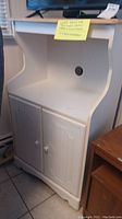 Left-front view of white wooden microwave stand with cabinet showing two paneled doors and curved top shelf with cable hole.