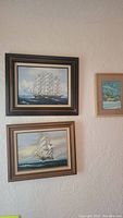 Three framed oil paintings hung on a white textured wall; two larger paintings of ships in darker wood frames and one smaller painting of a cabin in a lighter wood frame.