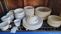 Complete view of the dinnerware set with coffee cups, saucers, large plates and dessert plates arranged on a shelf. Pieces are white with blue floral decoration.