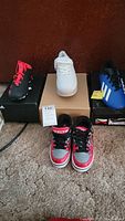 Four pairs of shoes: three pairs of cleats (black/red Adidas, white Adidas, blue Adidas) and one pair of black/red/grey Heelys with pink accents on front, shown on carpet.