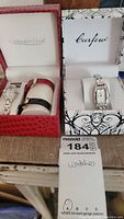 Two watches in their respective boxes: Coldwater Creek watch with red and black leather strap and Curfew watch with silver-tone metal bracelet.