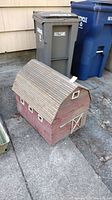 Wooden barn shaped yard decoration placed on pavement, showing its front and side angles with a textured shingle roof and small windows.