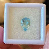 Close-up of pear-shaped sky blue topaz gemstone in white display box showing its facets and color.
