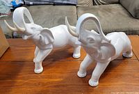 Two white ceramic elephant figurines displayed on wooden table, showing their size and condition.