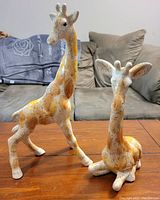 Photo showing two ceramic giraffe figurines on a wooden table with a gray sofa and blanket in background