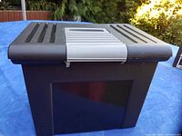Closed black hard plastic file box outdoors on a blue surface showing the top lid with ventilation slots.