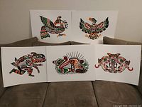 Photo showing five First Nations art prints by John Nelson displayed on a brown couch, featuring stylized traditional figures in green, red, black, and white colors including an Eagle, Thunderbird, Sea Monster, Sea Otter, and a Longhouse Mask.