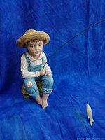 Full view of the boy fishing statue with fishing rod and fish separated, showing the entire figure sitting on a rock base against a blue background.