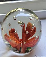 Clear glass orb paperweight showcasing the orange flower and embedded air bubbles inside the glass.