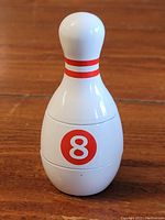 Front view of the herb grinder shaped like a bowling pin, showing white body with red stripes and an 8 ball logo.