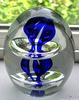 Egg-shaped clear glass paperweight with a cobalt blue swirl inside, shown standing upright against a window background.