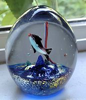 Front view of egg-shaped clear glass paperweight showing black and white fish inside with blue mounts and gold flecks on base.