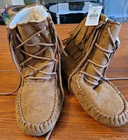 Photo showing pair of tan moccasin style boots with suede exterior, fuzzy white interior lining, fringe on sides, and laces.