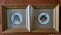 Pair of square wooden frames containing circular needlepoint portraits of Indigenous children with braided hair and feathers, showing wear on frames.