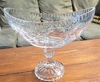 Clear crystal pedestal dish on wooden table with a sofa in background, showing detailed pinwheel and starburst cuts, no damages visible.