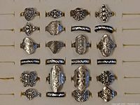 Photo showing assortment of 24 silver-tone light metal rings in a display box. Varied designs including butterfly, floral, geometric, filigree and plain bands with black enamel accents.