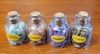 Photo showing four small corked glass bottles containing different semi-precious stones labeled Green Aventurine, Carnelian, Amethyst, and Gold Tiger's Eye, each approximately 1.5 inches tall.