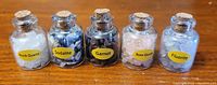 Photo of 5 small glass bottles each filled with different semi precious stones labeled as Rock Quartz, Sodalite, Garnet, Rose Quartz, and Fluorite sitting on a wood surface